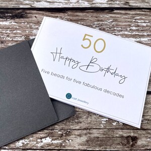 May include: A white card with gold lettering that says "50 Happy Birthday" and "Five beads for five fabulous decades". The card is on a wooden surface with a black envelope.