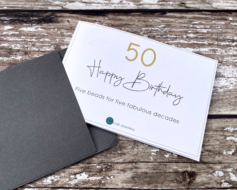 May include: A white card with gold lettering that says "50 Happy Birthday" and "Five beads for five fabulous decades". The card is on a wooden surface with a black envelope.