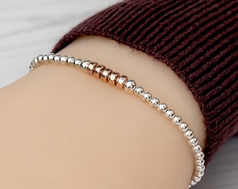 May include: A stretch bracelet composed of small, polished silver-toned spherical beads. The center of the bracelet features a sequence of four flat, rose gold-toned cylindrical spacer beads, creating a subtle contrast in color and shape.