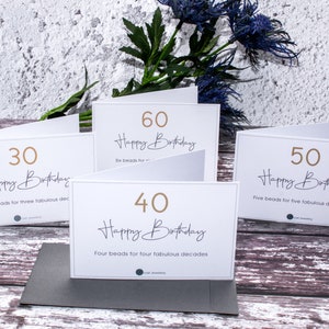 May include: Four white cards with gold numbers and text. The cards are arranged in a row on a wooden surface. The numbers on the cards are 30, 40, 50, and 60. The text on the cards says "Happy Birthday" and includes the number of beads for each age.