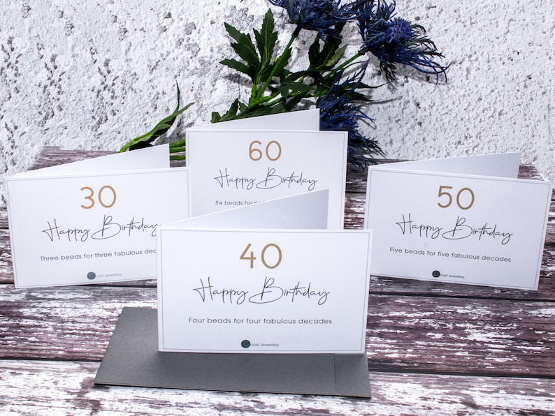 May include: Four white cards with gold numbers and text. The cards are arranged in a row on a wooden surface. The numbers on the cards are 30, 40, 50, and 60. The text on the cards says "Happy Birthday" and includes the number of beads for each age.