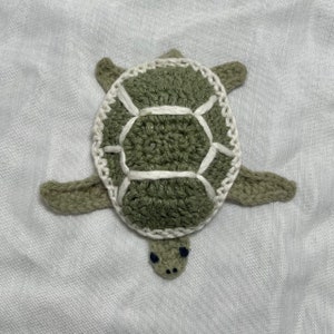 May include: A crocheted green turtle with a white shell and black eyes. The turtle is lying on its back.
