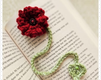 Puede incluir: Un marcapáginas de amapola roja de ganchillo con un tallo y hojas de ganchillo verde. El marcapáginas está colocado sobre un libro abierto.