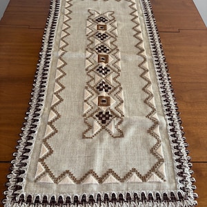 May include: Rectangular beige table runner with intricate brown and tan embroidered geometric patterns. The edges feature a decorative trim with a scalloped design. The runner is placed on a wooden table, showcasing its decorative appeal.