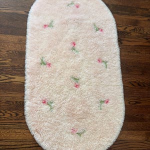 May include: An oval-shaped, light peach-colored rug with a high-pile, shaggy texture. The rug features a repeating pattern of small, embroidered pink flowers with green stems scattered across the surface in a soft, plush material.