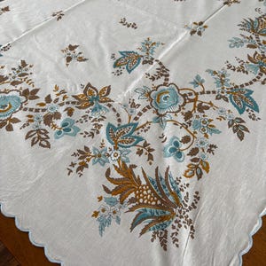 May include: White tablecloth with a floral pattern in teal and brown. The edges have a scalloped trim in light blue. The floral design includes stylized flowers and leaves, creating a vintage aesthetic.