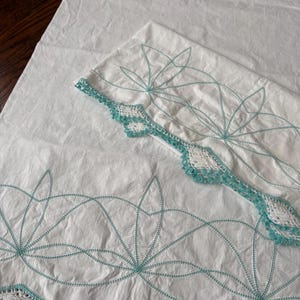 May include: White bed linens with a decorative design. The linens feature a teal embroidered floral pattern and a crocheted border. The fabric appears to be cotton or a similar material. The linens are folded on a wooden surface.