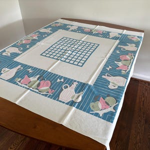 May include: A rectangular tablecloth featuring a central white grid pattern on a cream background. The border is a deep blue with white stripes, decorated with repeating illustrations of light pink pitchers and clusters of green and pink pears.
