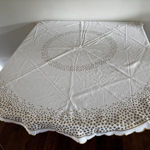 May include: A white tablecloth featuring a gold star pattern. The design includes a large central circular border of stars and a dense border of stars and small clouds along the scalloped edges. The fabric has a subtle, repeating geometric texture.