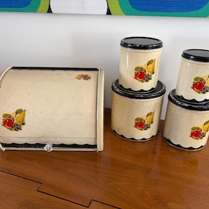May include: A vintage kitchen set featuring a bread box and four canisters. The off-white pieces have black trim and lids, with a fruit design of red apples, yellow pears, and purple grapes. The bread box has a roll-top lid and a crystal knob.