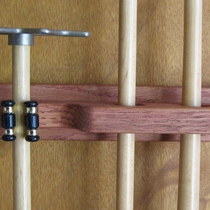 May include: A wooden wall mount for storing pool cues. The mount has a reddish-brown stain and features two black rubber holders for the cues. The mount is attached to a light brown wall.