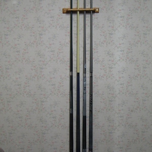 May include: A wooden wall mount with five pool cues. The cues are black, gray, and white. The wall mount is brown and has a simple design.