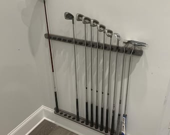 May include: A wall-mounted golf club rack with 14 clubs, including a driver, putter, and irons. The rack is made of black metal and has a gray finish. The clubs are arranged in order of size, with the driver at the top and the putter at the bottom.