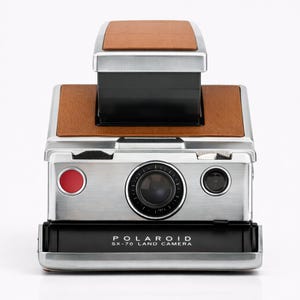Op de afbeelding: Een vintage Polaroid SX-70 Land Camera, met een geborstelde metalen behuizing en bruine lederen accenten. De camera heeft een prominente lens, een rode knop en de tekst "POLAROID SX-70 LAND CAMERA" op de voorkant. Een klassieke instant camera.