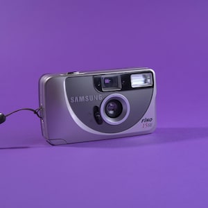 以下が含まれることがあります： シルバーのSamsung Fino 15SEコンパクトカメラ。内蔵フラッシュとリストストラップ付き。カメラの前面にはグレーのアクセントがあり、紫色の背景に配置されています。