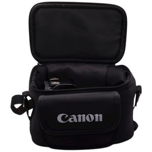Può includere: Borsa nera per fotocamera Canon con parte superiore aperta, che rivela l'interno. La borsa ha una tasca frontale con il logo Canon in bianco. È dotata di tracolla e maniglia. Progettata per proteggere e trasportare l'attrezzatura fotografica.