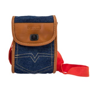 Puede incluir: Bolso bandolera Polaroid de mezclilla y cuero con correa roja. El bolso presenta una solapa de cuero marrón con el logotipo de Polaroid y un cierre negro. El cuerpo del bolso es de mezclilla azul con costuras amarillas.