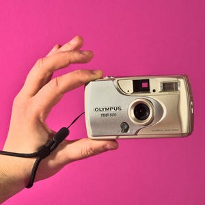 May include: Silver Olympus Trip 500 camera with a black wrist strap, held against a bright pink background. The camera has a lens, flash, and various buttons. The camera is a vintage film camera.