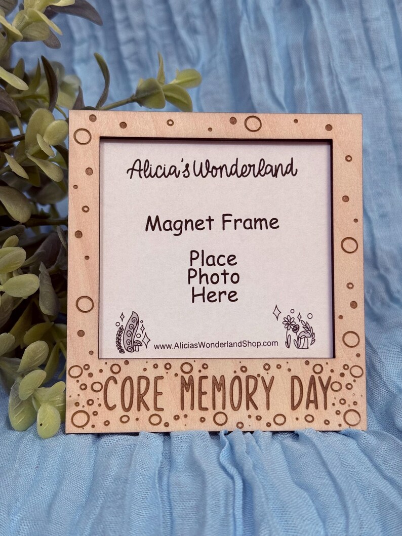 May include: A wooden magnet frame with the text "Alicia's Wonderland," "Magnet Frame," and "Place Photo Here." The frame has a light brown color with decorative circles and the words "CORE MEMORY DAY" at the bottom. The website address is also visible.