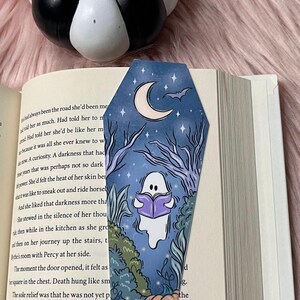 May include: A blue and purple coffin-shaped bookmark with a ghost reading a book, a crescent moon, a bat, and pumpkins. The bookmark has a starry night sky background.