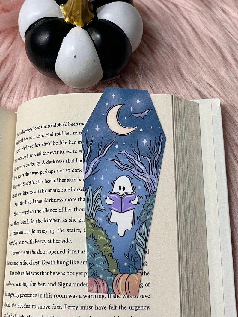 May include: A blue and purple coffin-shaped bookmark with a ghost reading a book, a crescent moon, a bat, and pumpkins. The bookmark has a starry night sky background.