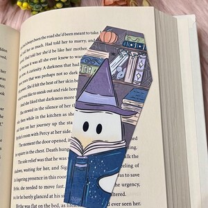 May include: A purple and blue book with a white, starry sky design. The book is being held by a witch with a black hat and a white face. The witch is standing in front of a bookshelf with books, a pumpkin, and a spider web.