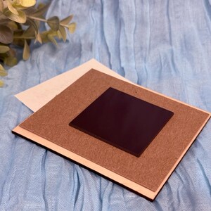 May include: A square magnet with a dark brown surface is centered on a light brown, rectangular backing. The backing is layered on a white rectangle, all set against a light blue fabric background. The magnet is likely for display or decorative purposes.