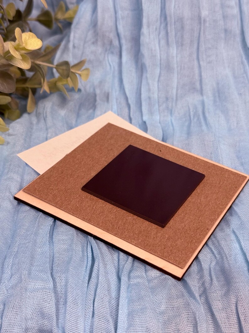 May include: A square magnet with a dark brown surface is centered on a light brown, rectangular backing. The backing is layered on a white rectangle, all set against a light blue fabric background. The magnet is likely for display or decorative purposes.