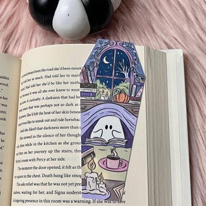 May include: A purple and blue coffin-shaped bookmark with a ghost reading a book, a cup of coffee, and two candles. The background shows a window with a crescent moon and a tree outside. The bookmark has the text "Death hung like smog" on it.