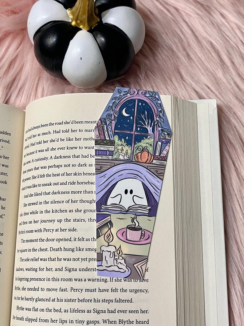 May include: A purple and blue coffin-shaped bookmark with a ghost reading a book, a cup of coffee, and two candles. The background shows a window with a crescent moon and a tree outside. The bookmark has the text "Death hung like smog" on it.