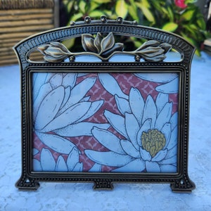 May include: A bronze-colored metal picture frame with a floral design. The frame holds a print with white flowers on a pink background. The top of the frame features a stylized flower and leaf design.