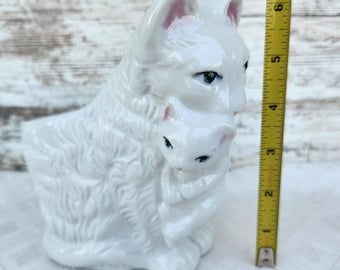 May include: White ceramic figurine of two cats, one larger cat holding a smaller cat in its arms. The cats have pink ears and black eyes.