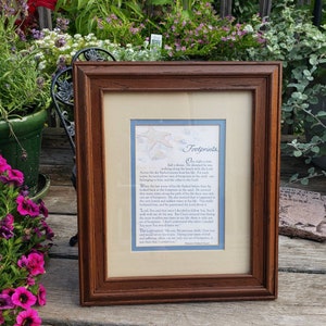 May include: A framed print with a brown wooden frame. The print features the poem "Footprints" with a light blue border and a beige background. The text is in a script font, and the image includes starfish.