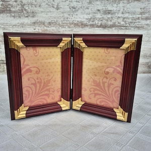 May include: A folding double photo frame in a dark red-brown hue, with gold-coloured corner detailing. The interior displays a peach-coloured patterned background. The frame is designed to stand upright.