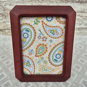 Peut inclure: Un cadre photo rectangulaire en bois rouge foncé. Le cadre contient une impression cachemire colorée avec des détails jaunes, verts, bleus et oranges. Le cadre a un design légèrement incliné.
