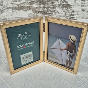 May include: A hinged, gold-colored photo frame with two panels. One panel displays the text "3 1/2 x 5 in" and "9 x 13cm" along with the "ACME FRAMES" logo. The other panel contains a photo of a child on a beach with a kite.