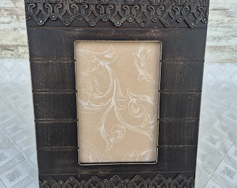 May include: A rectangular, dark brown picture frame with ornate, decorative metal trim. The frame has a rectangular opening with a beige background and a white floral design. The frame is made of wood and has a distressed finish.