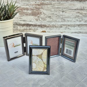 May include: A collection of five black metal photo frames in various styles. Some frames are hinged, and one displays the text "Alyssa". The frames are arranged on a white surface, with a potted plant in the background.