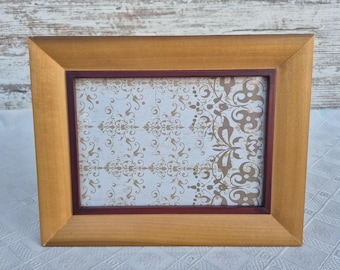 May include: A rectangular wooden picture frame with a light brown finish. The frame has a burgundy inner border and displays a patterned design in gold and brown on a white background. The frame is sitting on a white surface.