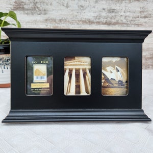 May include: Black picture frame with three openings. The frame has a raised edge and a smooth finish. The top opening has a price tag that reads "2x3 $19.99". The middle opening shows a black and white photo of a building with columns. The bottom opening shows a color photo of the Sydney Opera House.