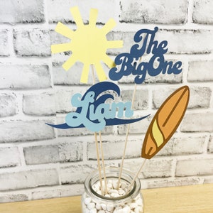 May include: A set of four paper party decorations for a first birthday party. The decorations include a yellow sun, a blue wave with the name Liam, a blue surfboard with the text "The Big One", and an orange surfboard. The decorations are on wooden skewers and are in a glass jar with white rocks.