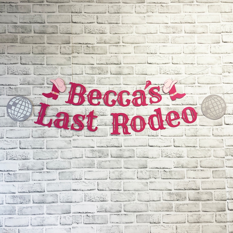 以下が含まれることがあります： 白いレンガの壁に、「Becca's Last Rodeo」と書かれたピンクと赤の横断幕。横断幕には、ピンクのウエスタンハットとブーツが描かれている。