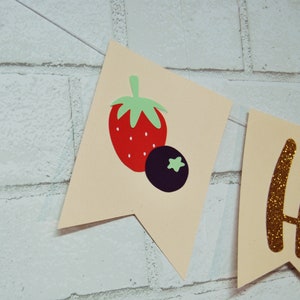 May include: A beige banner with a red and green strawberry and a purple blueberry. The banner also has a gold glitter letter "H" on it.