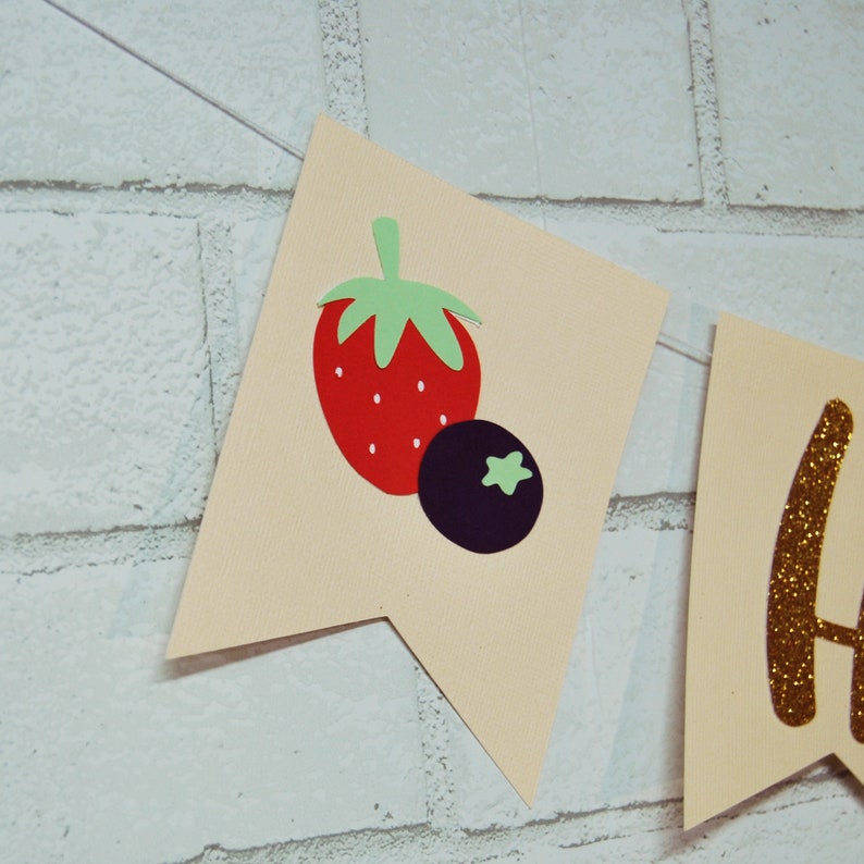 May include: A beige banner with a red and green strawberry and a purple blueberry. The banner also has a gold glitter letter "H" on it.