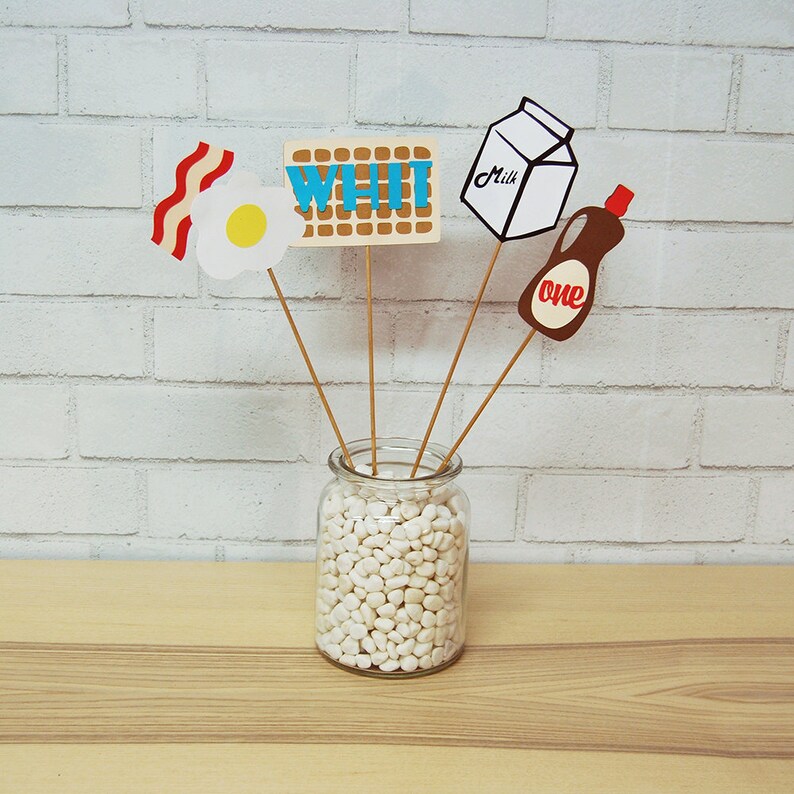 May include: A glass jar filled with white pebbles, with five paper cutouts on sticks inserted into the jar. The cutouts are a waffle with the word "WHI" on it, a carton of milk with the word "Milk" on it, a fried egg, a strip of bacon, and a bottle of syrup with the word "one" on it.
