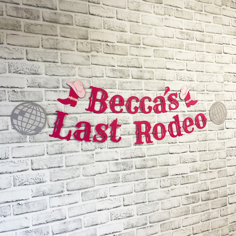 以下が含まれることがあります： 白いレンガの壁に、「Becca's Last Rodeo」と書かれた赤と白の横断幕がかけられています。横断幕には、ピンクのテンガロンハットが2つと、銀色のディスコボールが2つ付いています。
