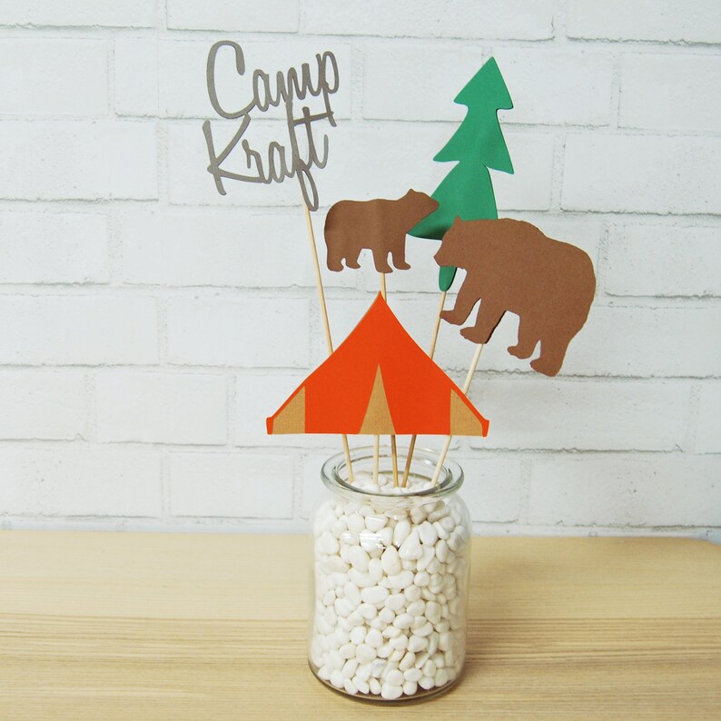 May include: A centerpiece featuring a red and brown tent, two brown bears, a green tree, and the words "Camp Kraft" in silver lettering. The items are arranged in a glass jar filled with white stones.