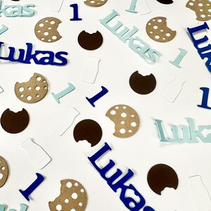 May include: Blue, brown and white confetti with the name "Lukas" and the number "1" scattered on a white background. The confetti is shaped like cookies, milk bottles and letters.