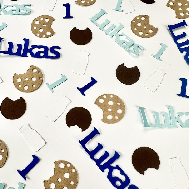 May include: Blue, brown and white confetti with the name "Lukas" and the number "1" scattered on a white background. The confetti is shaped like cookies, milk bottles and letters.