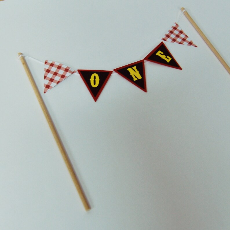 May include: Red and white gingham cake topper with the word "ONE" in yellow letters on black flags.